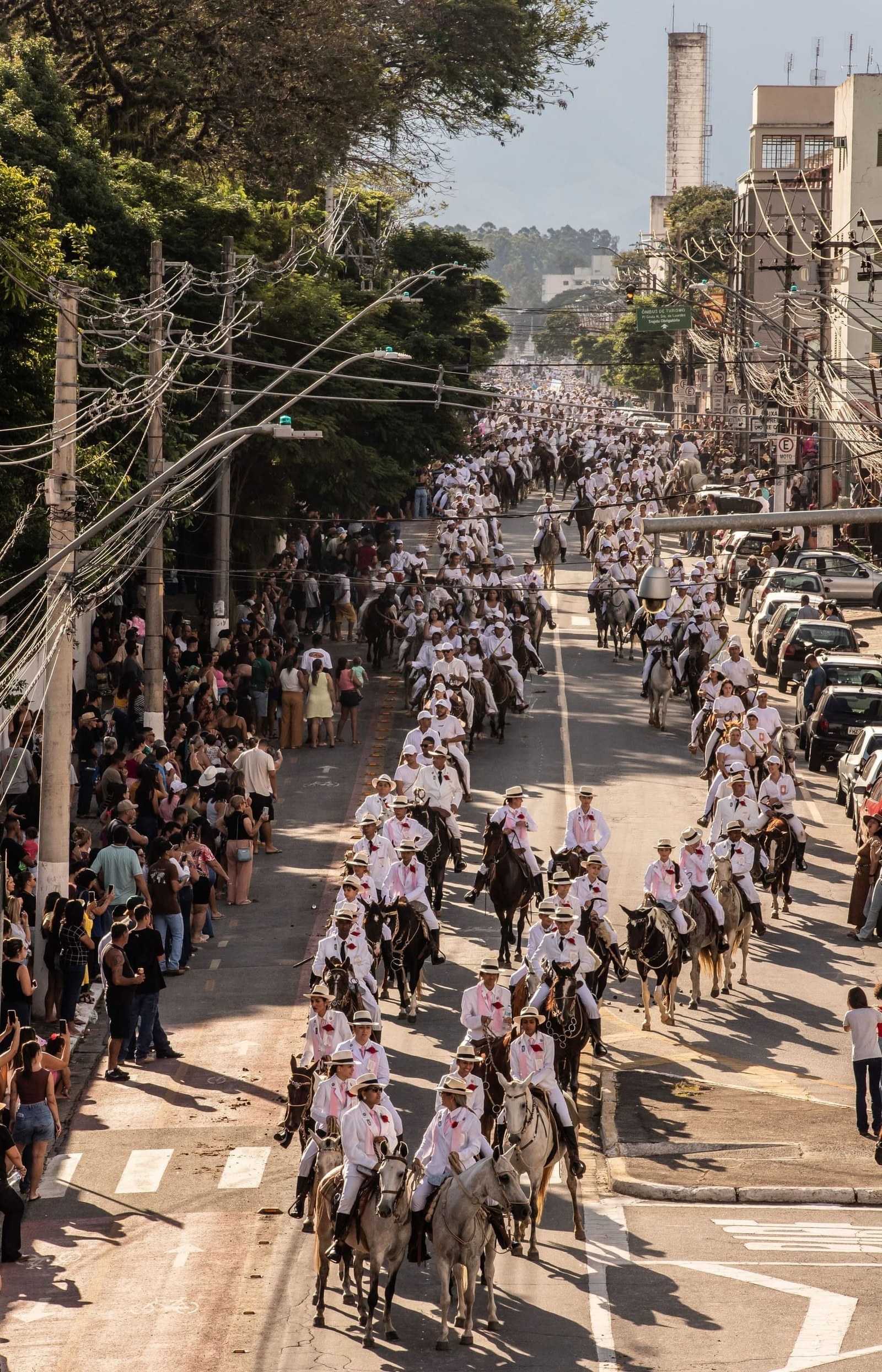 Do alto, a longa fila de cavaleiros de branco ocupa a avenida, com o público nas calçadas
