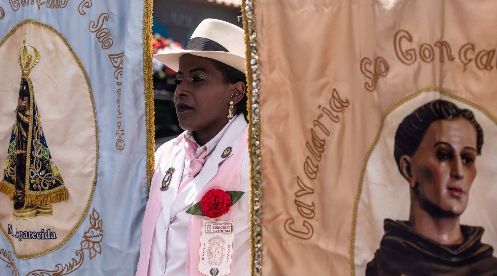 Uma Mantena de gravata e faixa cor-de-rosa e cravo vermelho entre os estandartes de Nossa Senhora Aparecida e de São Gonçalo