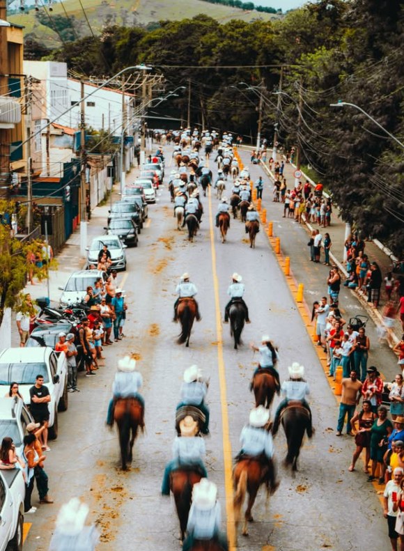 Vista do alto: dezenas de cavaleiros de chapéu branco descem a rua em fila, com o público nas calçadas